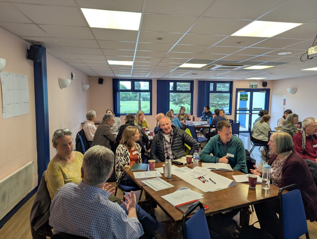 A wide shot of a community workshop held in a bright room with large windows. Multiple groups of people are seated around wooden tables covered in notes and flipchart paper, engaged in active discussion.
