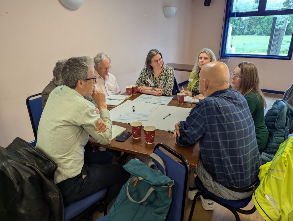 To make the alt text more specific to your workshop in Gloucestershire, you can weave in the actual event details. This is great for SEO and for providing clear context to anyone using a screen reader. Alt Text for the wide shot: "Participants at the Gloucestershire Food Systems Ideas Exchange workshop engaged in group discussions at wooden tables, collaborating on local food strategy and system change.