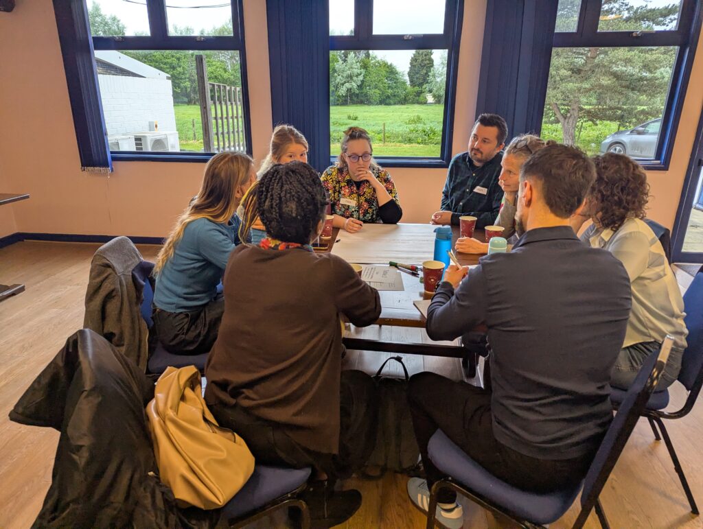 To make the alt text more specific to your workshop in Gloucestershire, you can weave in the actual event details. This is great for SEO and for providing clear context to anyone using a screen reader. Alt Text for the wide shot: "Participants at the Gloucestershire Food Systems Ideas Exchange workshop engaged in group discussions at wooden tables, collaborating on local food strategy and system change.